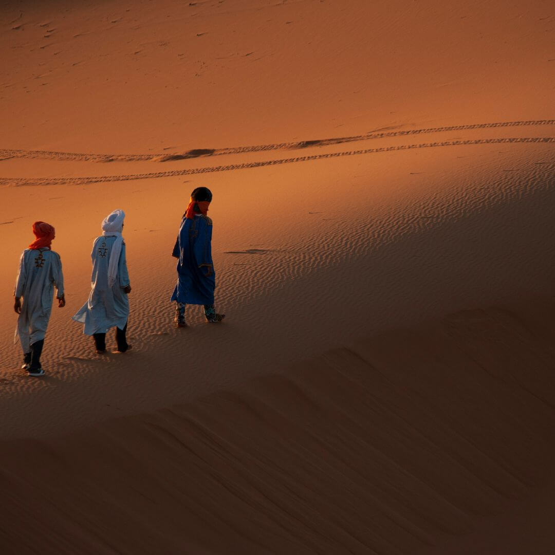صورة لقافلة جمال تسير ببطء فوق الكثبان الرملية الذهبية في قلب صحراء مرزوكة تحت سماء صافية زرقاء
